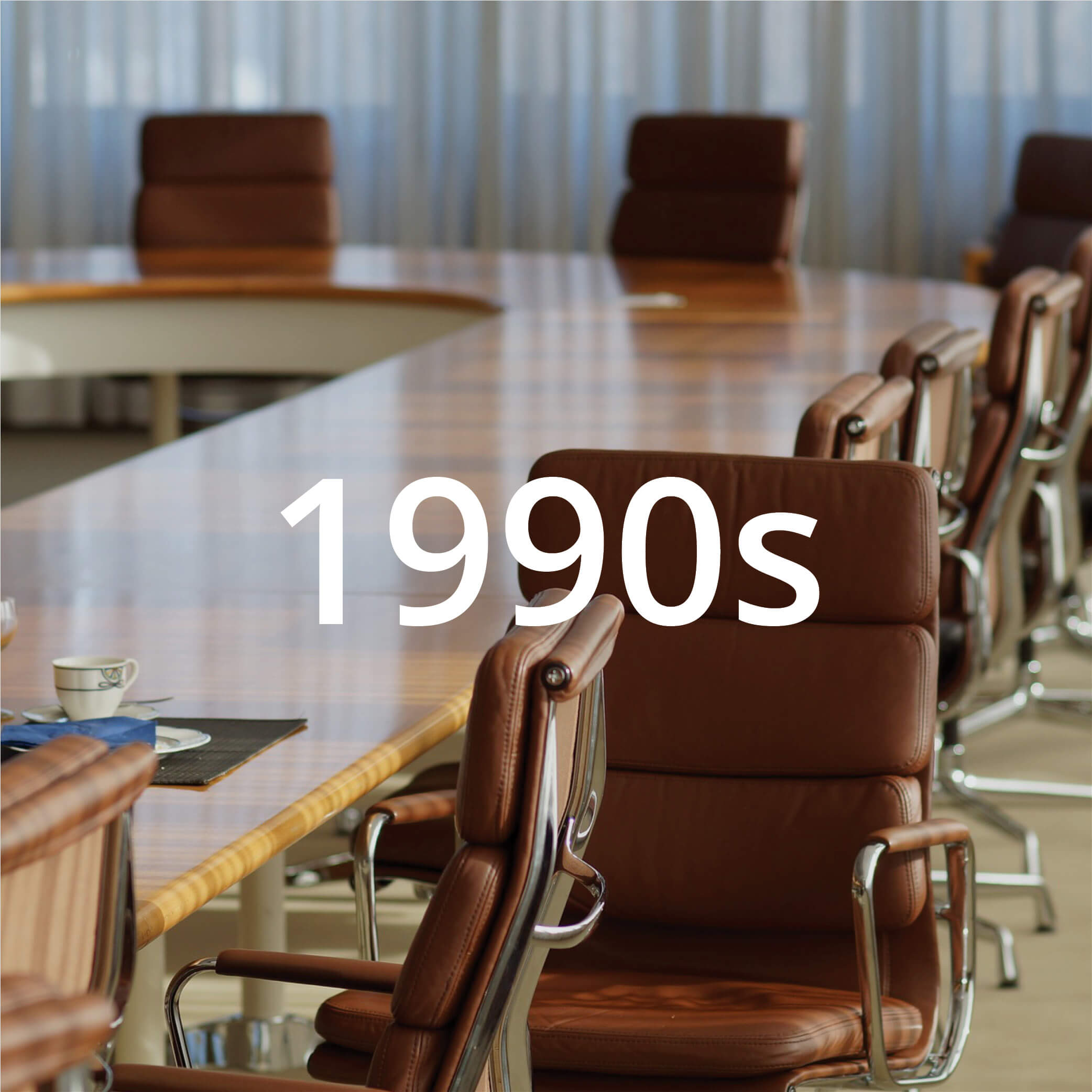 Photo of a meeting room featuring a u-shaped table and modern brown leather chairs. Photo also features text overlay that reads "1990s".